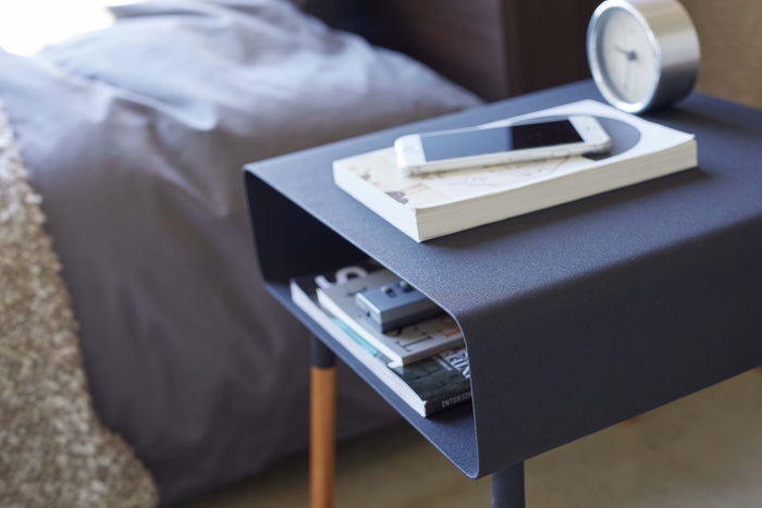 Storage Table from Yamazaki Home being used as a nightstand in a modern bedroom. Shows interior storage capabilities and slim design.  