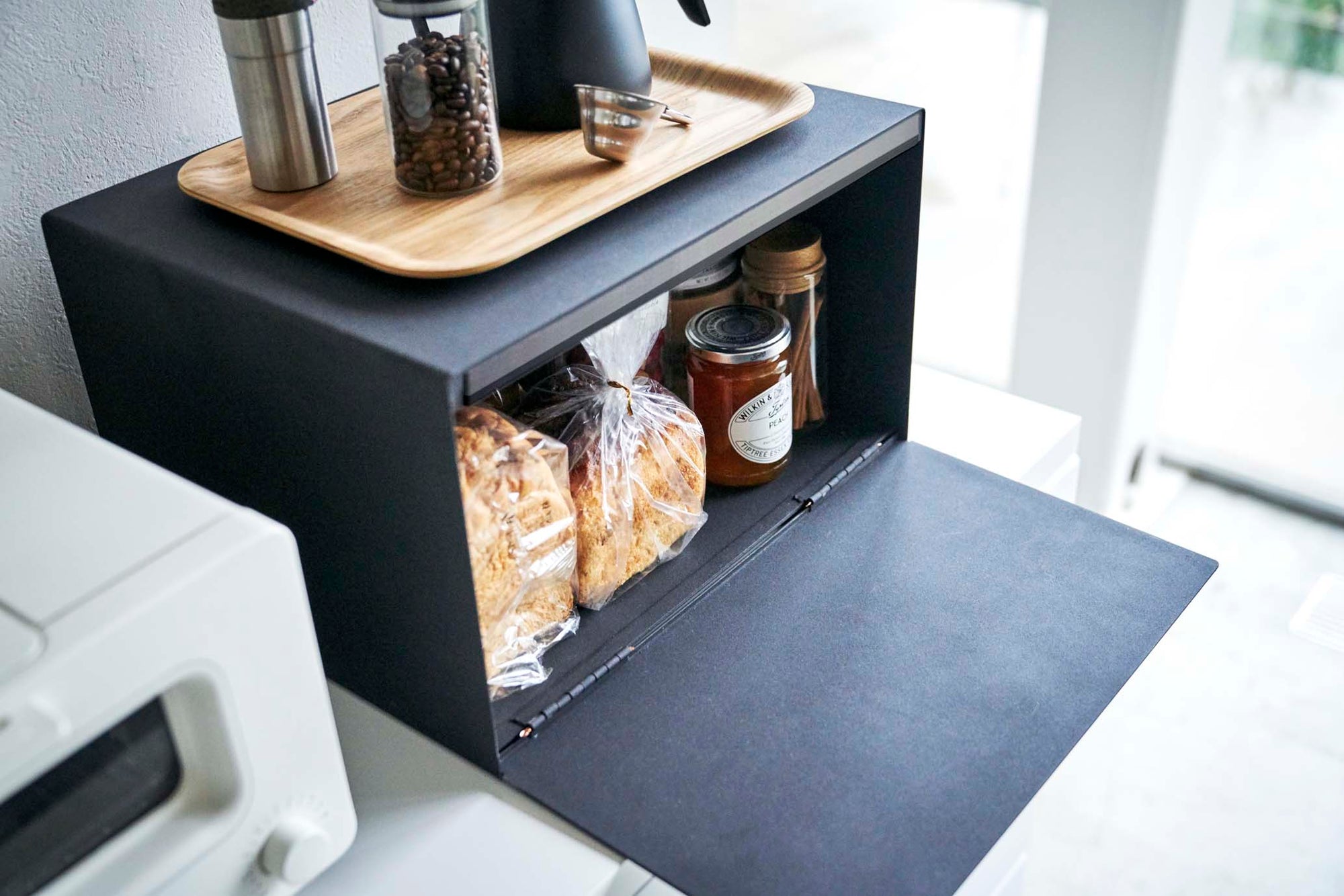 Bread Box from Yamazaki Home loaded with pastries, jam, snacks, and breads in a modern kitchen with coffee supplies on top. The black is striking against all-white kitchen. 