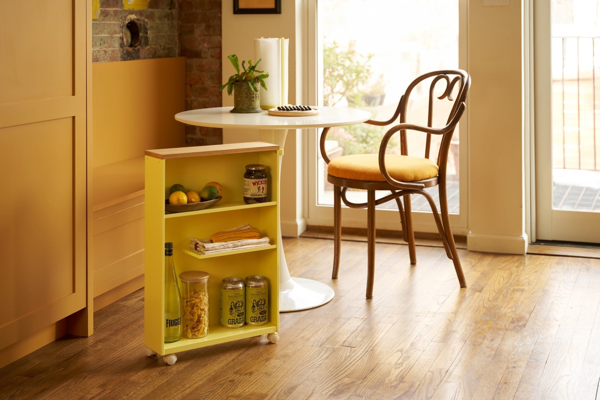 Yellow rolling storage cart holding pantry items in a warm kitchen with wood floor. Cafe table and chair in the background set as breakfast nook. 