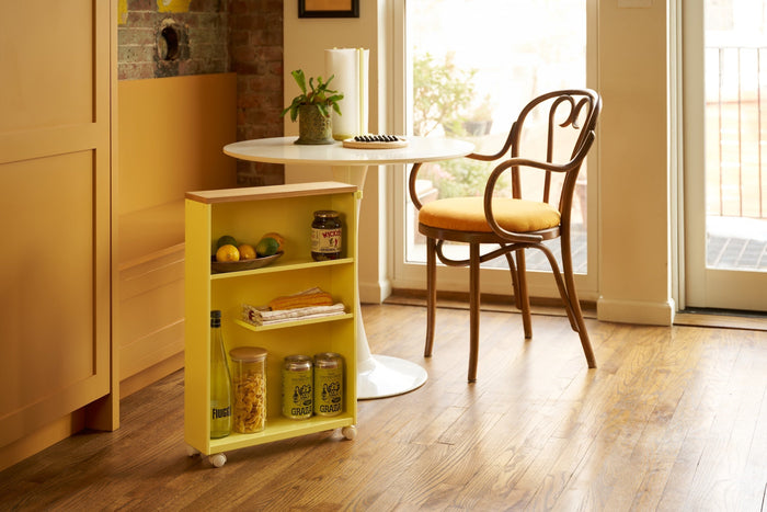 Yellow rolling storage cart holding pantry items in a warm kitchen with wood floor. Cafe table and chair in the background set as breakfast nook. 