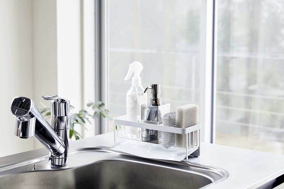 White steel sponge and bottle organizer sitting on a modern steel sink in a light-filled kitchen.