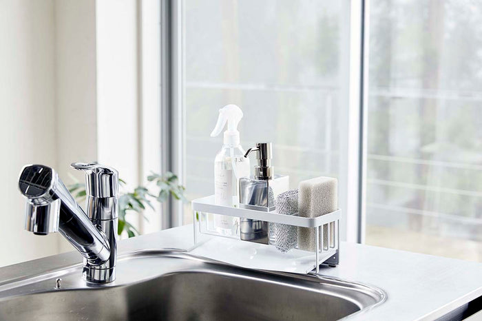 White steel sponge and bottle organizer sitting on a modern steel sink in a light-filled kitchen.