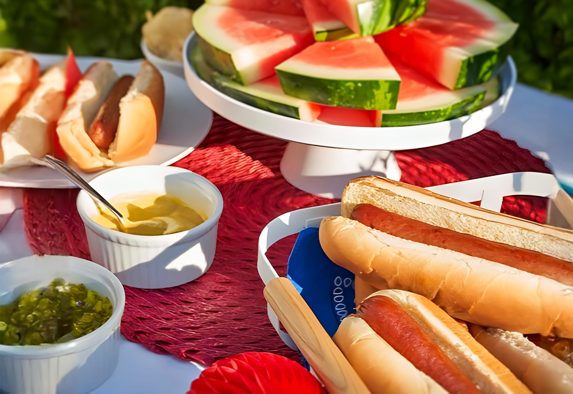 Picnic spread out with watermelon on a Yamazaki cake stand and hot dogs in a yamazaki fruit basket. There is a cup of dill and a cup of mustard. The table is decorated for Fourth of July in red, white and blue.