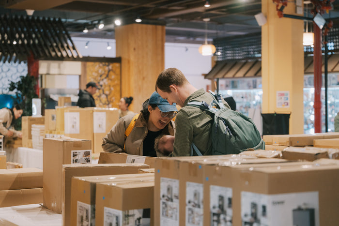 A man and woman are smiling at baby while standing amongst the rows of boxes at the Yamazaki Home Open Box Sale at Japan Village.
