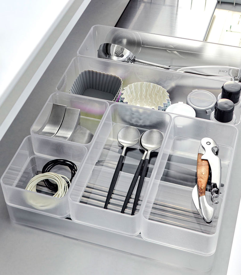 Close-up image of four different sizes of stackable clear organizers—small, medium, large, and deep—neatly placed in a bathroom drawer. Made from transparent material, they accommodate items like brushes and sanitary products, showcasing their clarity and stackability.