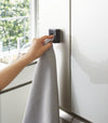 Black Yamazaki Home Traceless Adhesive Towel Holder attached to a cabinet with a towel being inserted