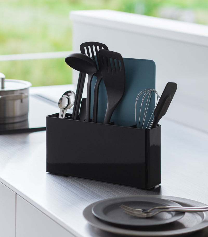 Black Utensil & Thin Cutting Board Holder by Yamazaki Home on a kitchen counter, holding various utensils.