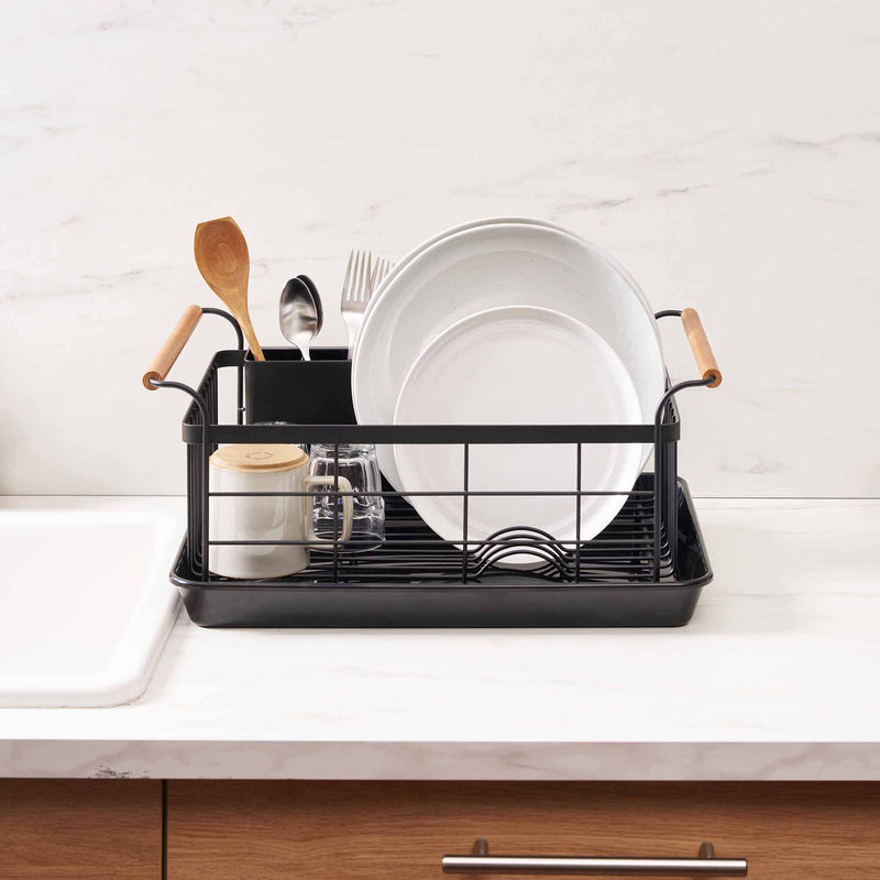 Black steel dish drying rack with wood handles on a drying tray in a modern kitchen with marble countertop near a sink from Yamazaki Home.