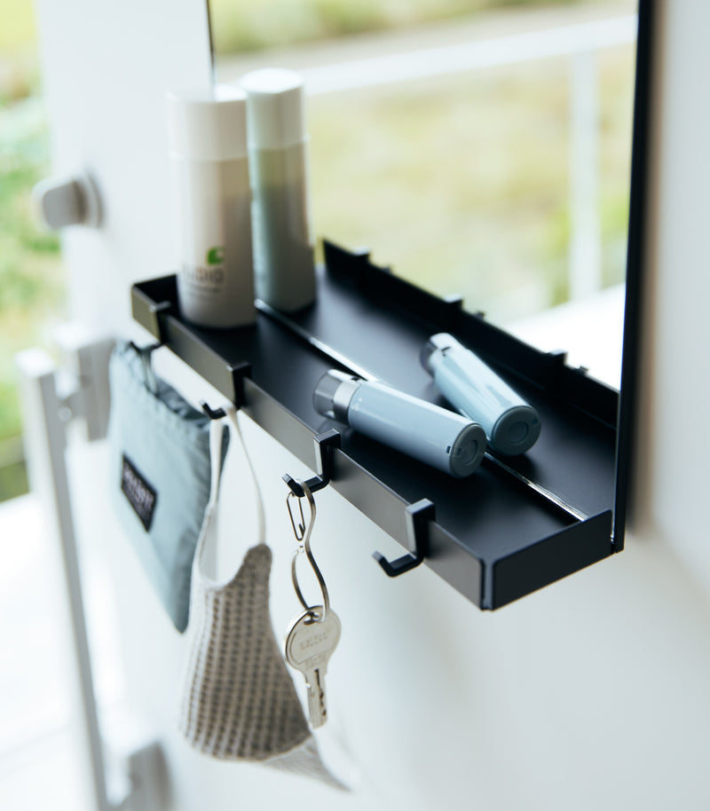 A close up of a black Yamazaki Magnetic Mirror With Storage Rack attached to a door with a hanging bag, mask, and key.