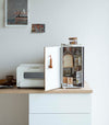 A vertical white metal breadbox is seen on a wooden kitchen counter next to a white microwave oven. The breadbox’s door is swung open to the left and bread, other grains, and a bottle of wine are seen inside the box. A magnetic stop is seen opposite the open door. On the open door is a white hook holding brown plastic measurement spoons. On top of the box is a folded towel and plastic container of cookies.