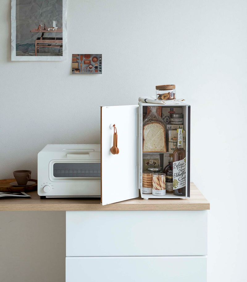 A vertical white metal breadbox is seen on a wooden kitchen counter next to a white microwave oven. The breadbox’s door is swung open to the left and bread, other grains, and a bottle of wine are seen inside the box. A magnetic stop is seen opposite the open door. On the open door is a white hook holding brown plastic measurement spoons. On top of the box is a folded towel and plastic container of cookies.