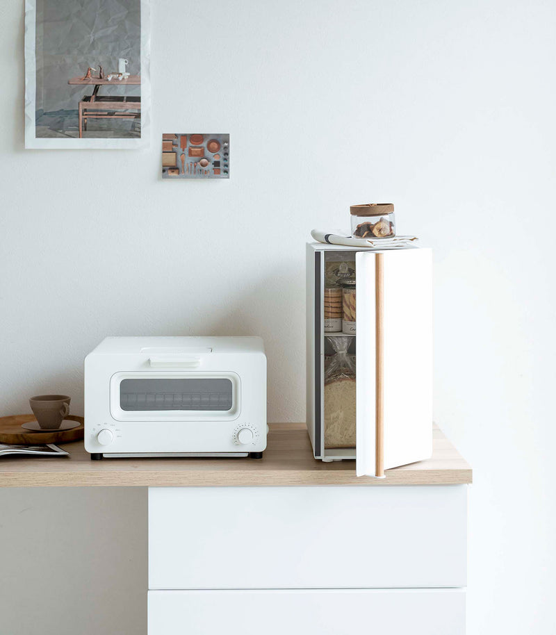 A vertical white metal breadbox, with a vertical wooden handle, is seen on a white kitchen counter next to a white microwave oven and a wooden plate with a brown coffee cup and matching saucer on it.