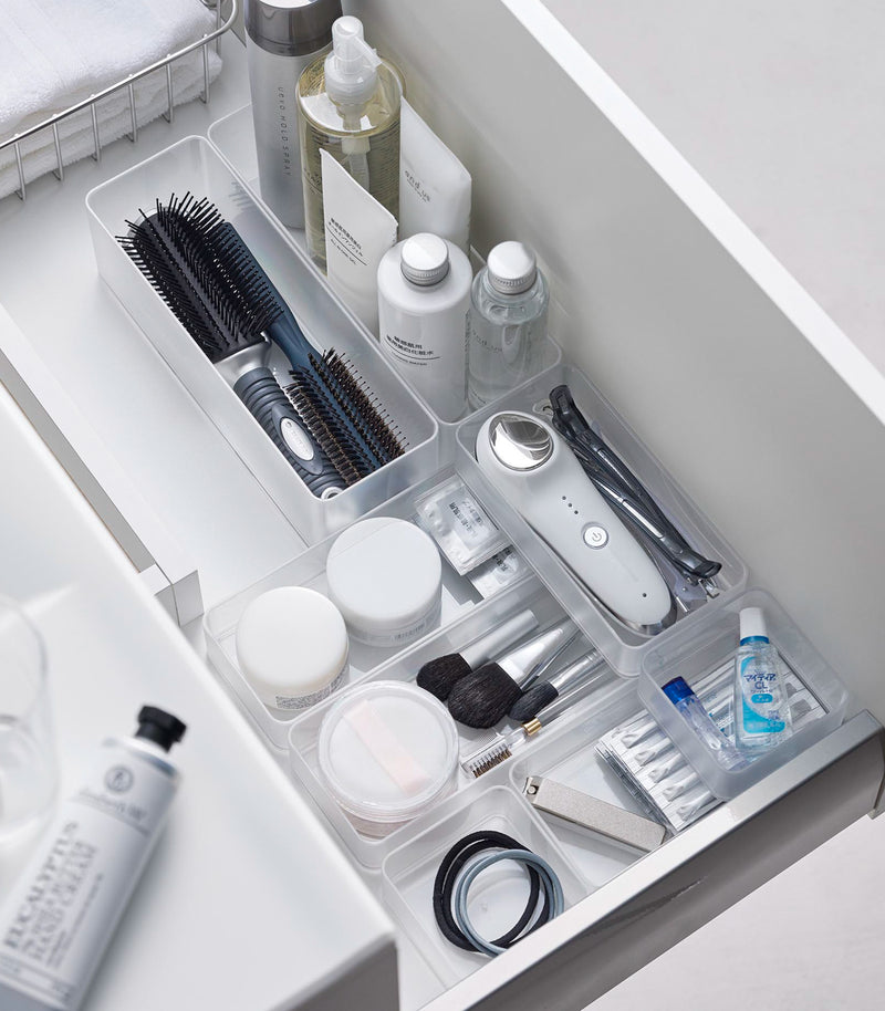 Four different sizes of stackable clear organizers—small, medium, large, and deep—are neatly placed in a bathroom drawer. Made from transparent material, they accommodate items like brushes and sanitary products, showcasing their clarity and stackability.