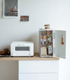 A vertical white metal breadbox is seen on a wooden kitchen counter next to a white microwave oven. The breadbox’s door is swung open to the right and bread, other grains, and a bottle of wine are seen inside the box. A magnetic stop is seen opposite the open door. On the open door is a white hook holding brown plastic measurement spoons. On top of the box is a folded towel and plastic container of cookies.