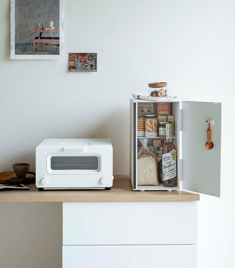 A vertical white metal breadbox is seen on a wooden kitchen counter next to a white microwave oven. The breadbox’s door is swung open to the right and bread, other grains, and a bottle of wine are seen inside the box. A magnetic stop is seen opposite the open door. On the open door is a white hook holding brown plastic measurement spoons. On top of the box is a folded towel and plastic container of cookies.