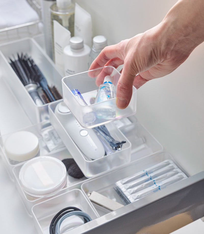 Four different sizes of stackable clear organizers—small, medium, large, and deep—are neatly placed in a bathroom drawer. Made from transparent material, they accommodate items like brushes and sanitary products, showcasing their clarity and stackability.