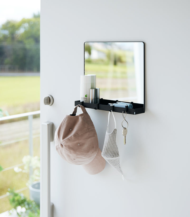 A black Yamazaki Magnetic Mirror With Storage Rack attached to a door with a hanging hat, mask, and key with a view outside.