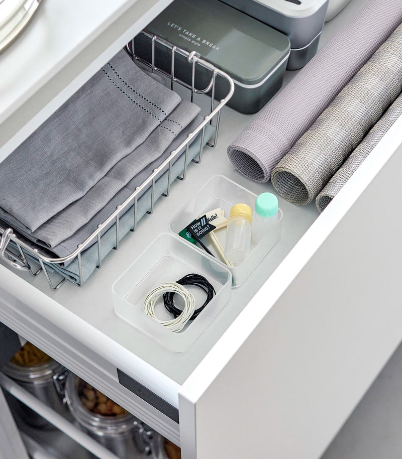 Two small stackable clear organizers are placed in a kitchen drawer, organizing small items like strings and tiny sauce bottles