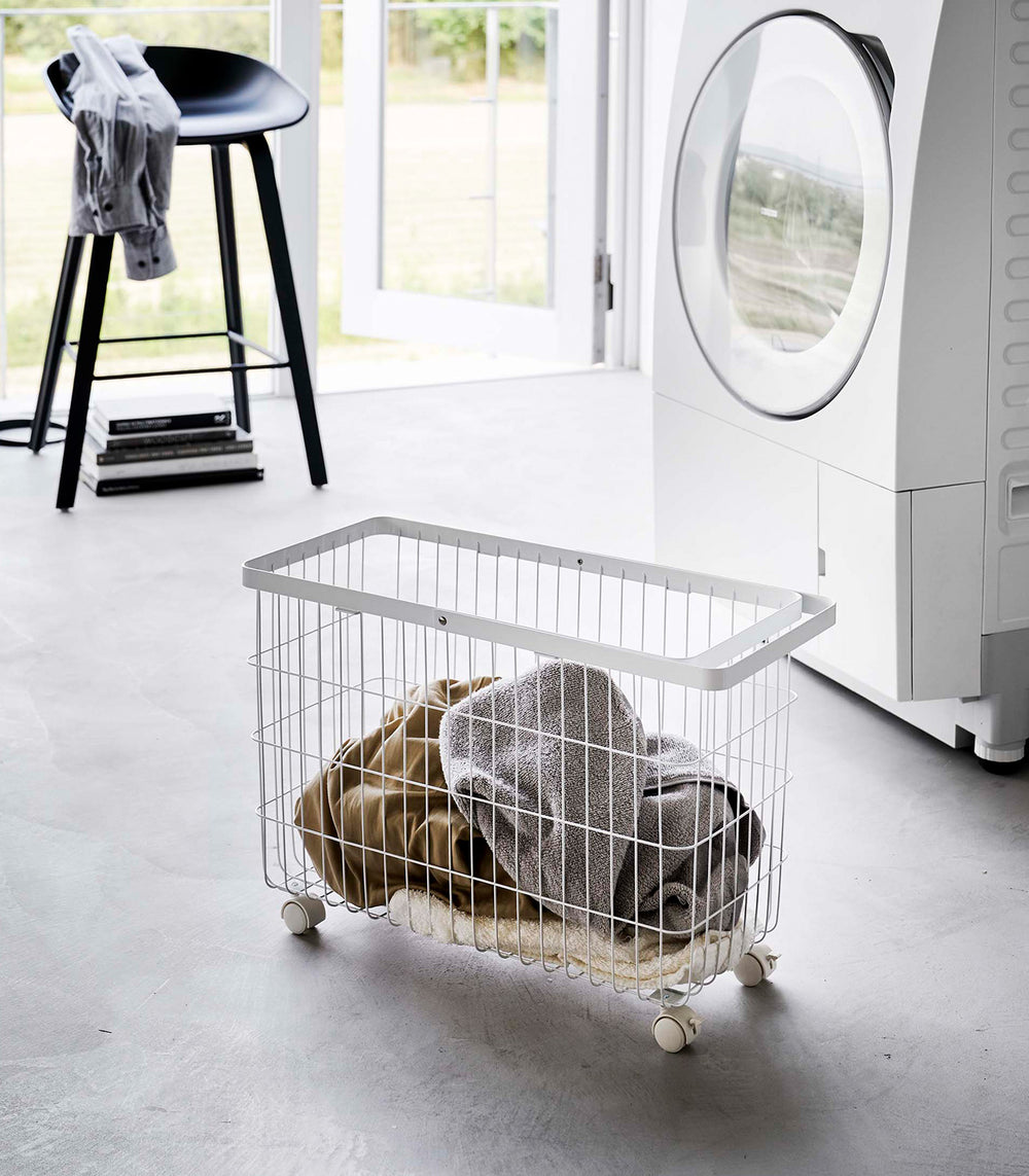 Rolling Wire Basket by Yamazaki Home in white in a sunny laundry room with several towels inside.