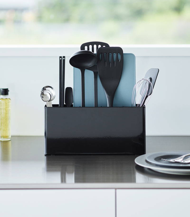 Black Utensil & Thin Cutting Board Holder by Yamazaki Home on a kitchen counter, holding various utensils and a cutting board.
