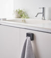 Black Yamazaki Home Traceless Adhesive Towel Holder attached to a cabinet under a sink with a towel inserted