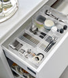 Four different sizes of stackable clear organizers—small, medium, large, and deep—are neatly placed in a kitchen drawer. They effectively organize various items such as cutlery, bottle openers, and small trays, showcasing their versatility and stackability
