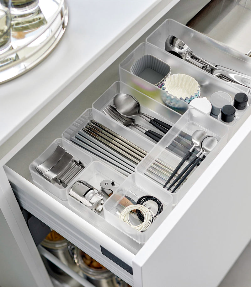 Four different sizes of stackable clear organizers—small, medium, large, and deep—are neatly placed in a kitchen drawer. They effectively organize various items such as cutlery, bottle openers, and small trays, showcasing their versatility and stackability