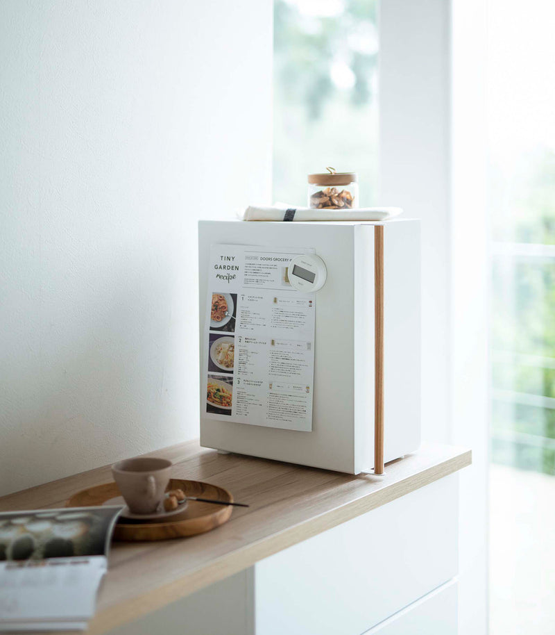 A profile view of a vertical white metal bread box sat on a wooden kitchen counter.  A long vertical wooden handle is positioned on the left at the front of the box. A magnetic timer is holding up a recipe card on the side of the breadbox. A folded towel and glass container of treats is laid on top. On the counter in next to the breadbox is a wooden plate with a brown coffee cup and matching saucer almost out of focus. Next to the wooden plate is an opened magazine.