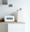 A vertical white metal breadbox, with a vertical wooden handle, is seen on a white kitchen counter next to a white microwave oven and a wooden plate with a brown coffee cup and matching saucer on it. On top of the breadbox is a folded towel and glass container of snacks. Behind the counter is a white wall with two different sized postcards positioned diagonally from each other.