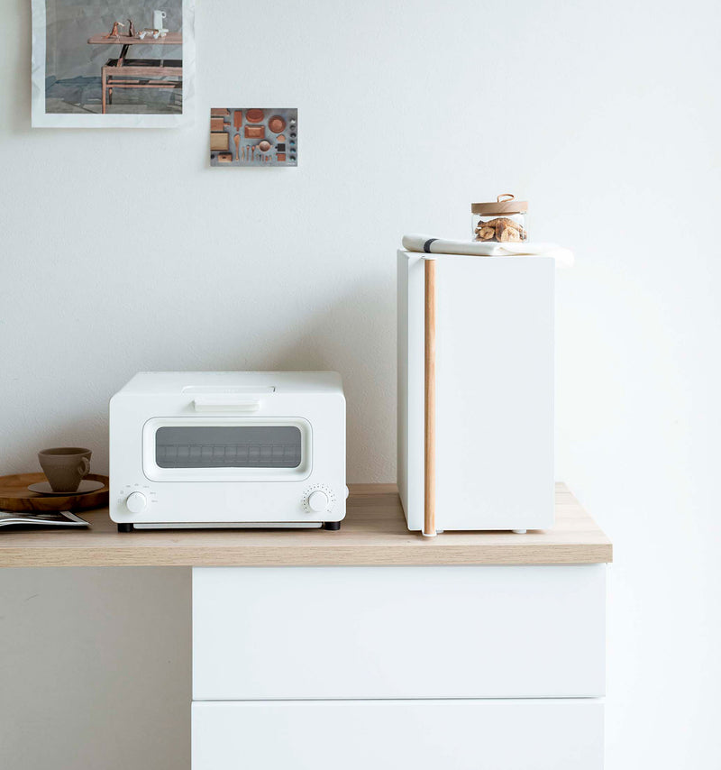 A vertical white metal breadbox, with a vertical wooden handle, is seen on a white kitchen counter next to a white microwave oven and a wooden plate with a brown coffee cup and matching saucer on it. On top of the breadbox is a folded towel and glass container of snacks. Behind the counter is a white wall with two different sized postcards positioned diagonally from each other.