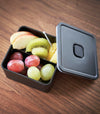 Close up aerial view of black Vacuum-Sealing Bento Box with cover ajar holding fruit by Yamazaki Home.