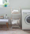 Front view of white Laundry Wagon and Basket holding clothes and towels in laundry room by Yamazaki Home.