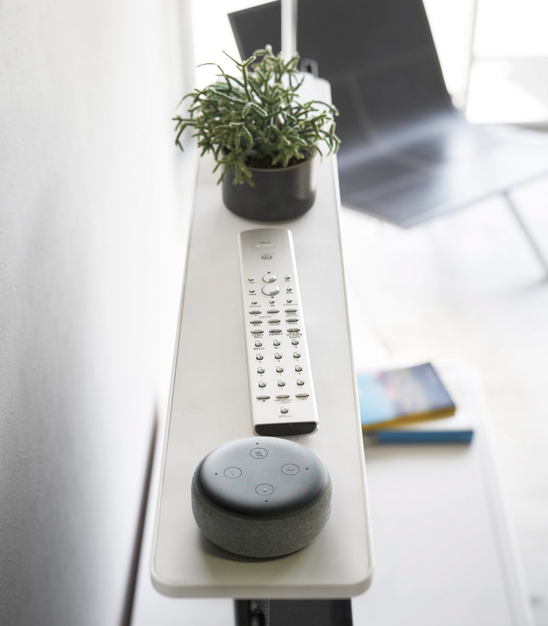 Side view of white Compliant TV Shelf holding smart home device, plant, and remote by Yamazaki Home.