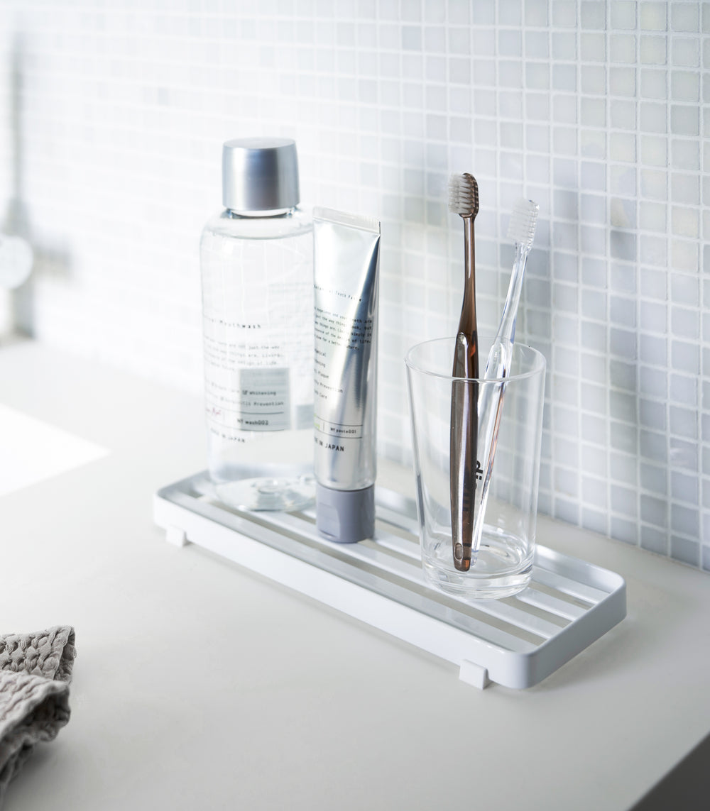 White slotted tray holding toothbrush and mouthcare items on bathroom sink counter by Yamazaki Home.