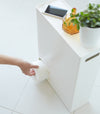 Aerial view of white Toilet Paper Stocker displaying phone, jar, and plant on top and dispensing a toilet paper roll by Yamazaki Home.